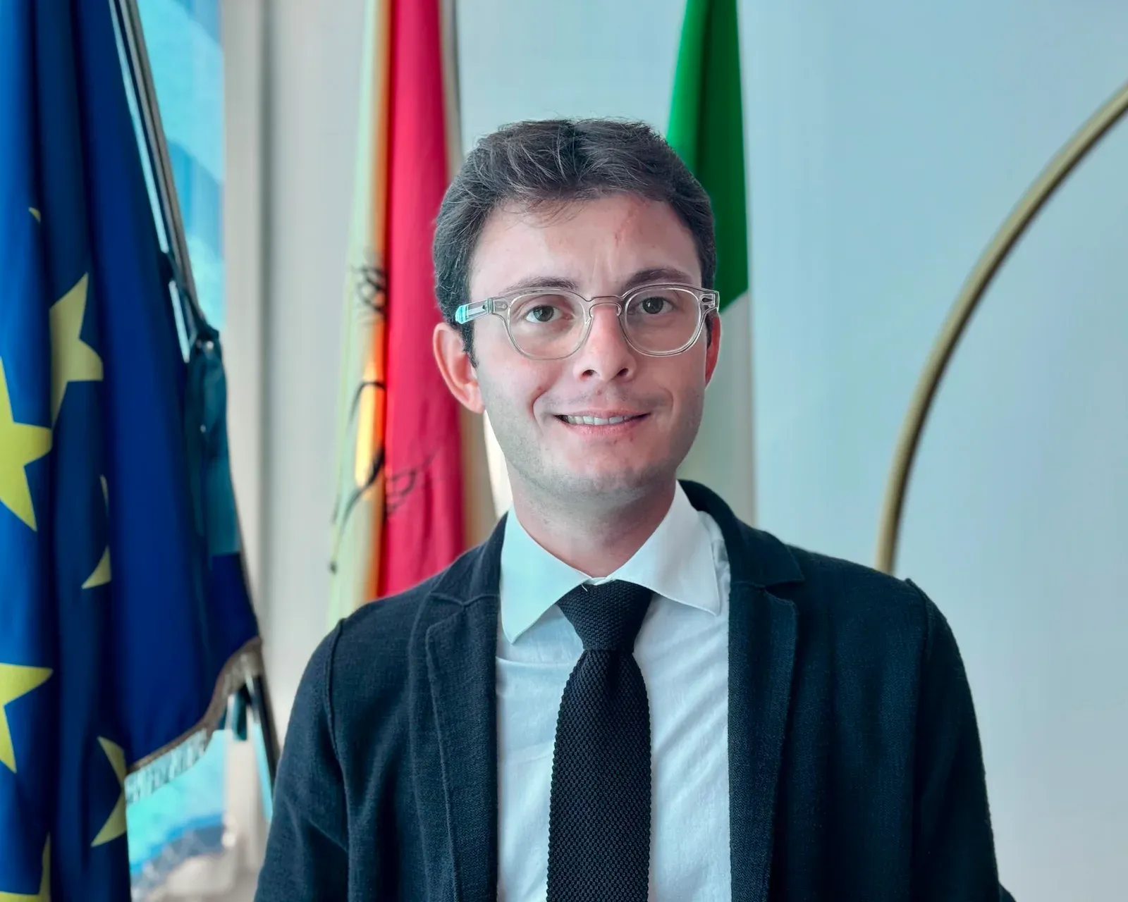 Luigi Genovese in giacca e cravatta, accanto alle bandiere.